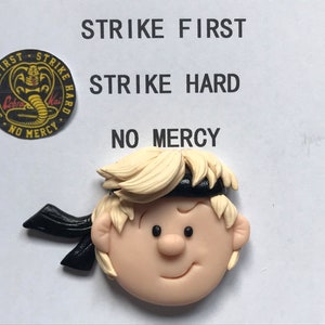 Puede incluir: Una pequeña figura artesanal de un niño pequeño con cabello rubio que lleva una cinta para la cabeza negra. La figura está hecha de arcilla polimérica y tiene un estilo de dibujos animados. El fondo es blanco con el texto "Strike First, Strike Hard, No Mercy" en negro.