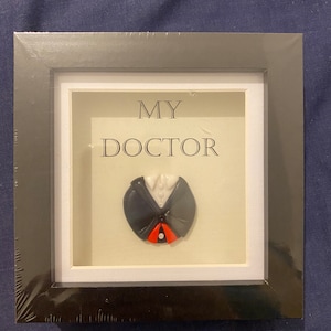 Può includere: Un'immagine incorniciata in nero con un passepartout bianco. L'immagine presenta una scultura in argilla 3D di un abito da medico con una cravatta rossa. Il testo "MY DOCTOR" è stampato sul passepartout.