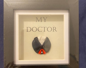 May include: A black framed picture with a white mat. The picture features a 3D clay sculpture of a doctor's outfit with a red tie. The text "MY DOCTOR" is printed on the mat.