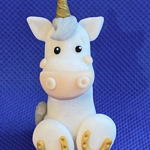 May include: A white unicorn figurine with a gold horn and hooves. The unicorn is sitting with its legs crossed and has a sparkly finish.