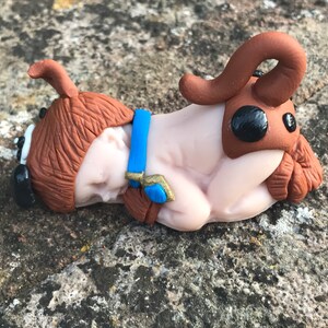 Könnte beinhalten: Eine kleine, schlafende Babyfigur aus braunem Ton, die ein braunes und blaues Kostüm mit einem gehörnten Kopfschmuck trägt.