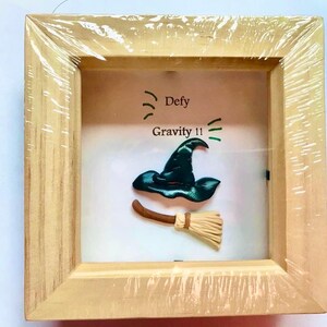 May include: A wooden frame containing a green witch's hat and a brown broom. The text "Defy Gravity!!" is written on the white background.