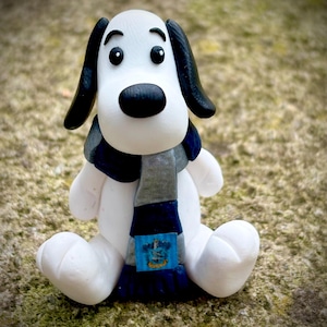May include: A white and black dog figurine with a black nose and ears, wearing a gray and navy blue scarf. The scarf has a blue emblem. The dog is sitting on a textured surface.