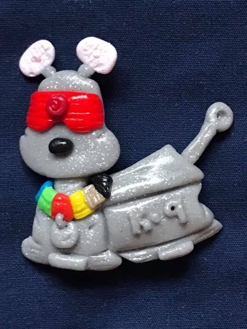 Peut inclure: Un jouet robot gris et paillet&eacute; avec des yeux rouges, des oreilles blanches et un bandeau color&eacute; autour du bras. Le robot tient un pinceau et porte l'inscription "K.I.Q" sur le c&ocirc;t&eacute;.