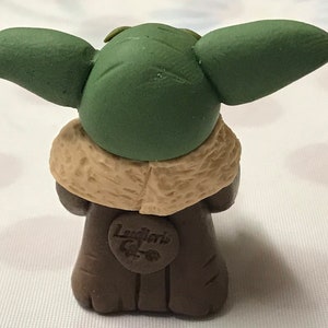 May include: A small green and brown figurine of a character from the Star Wars franchise. The figurine is wearing a brown coat with a tan fur collar. The figurine has large green ears and a small face. The figurine has a label on the front that says "Laughlin's Creations".