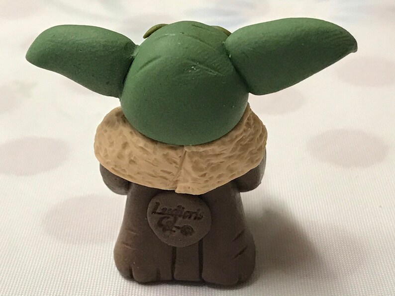 May include: A small green and brown figurine of a character from the Star Wars franchise. The figurine is wearing a brown coat with a tan fur collar. The figurine has large green ears and a small face. The figurine has a label on the front that says "Laughlin's Creations".