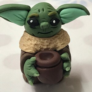 May include: A green and brown handmade figurine of a character from the Star Wars franchise, holding a brown pot in its hands.