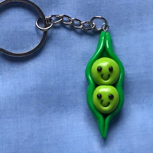May include: A green pea pod keychain with two smiling green peas inside. The keychain has a silver metal ring.