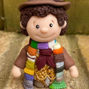 May include: A brown and white figurine of a person wearing a brown hat, a brown jacket, a colourful scarf, and a brown bow tie. The figurine has brown hair and a smiling face.