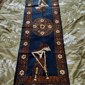 Peut inclure: Un chemin de table rectangulaire en tissu présentant un fond bleu profond avec des motifs complexes de style batik en brun et blanc. Le design comprend deux silhouettes de marionnettes d'ombre et un motif floral central circulaire.