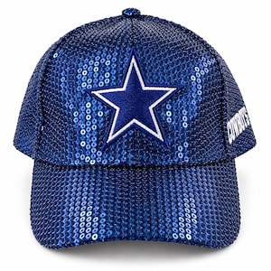 Puede incluir: Una gorra de béisbol azul real completamente cubierta de lentejuelas brillantes. El panel frontal presenta una gran estrella de cinco puntas bordada con un borde blanco. La palabra COWBOYS está bordada en texto blanco en el lateral.