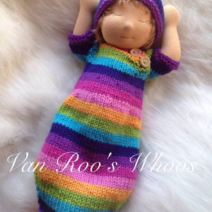 Puede incluir: Una muñeca de punto hecha a mano con un saco de dormir a rayas y un gorro morado y azul. La muñeca tiene los ojos cerrados y una expresión dulce.