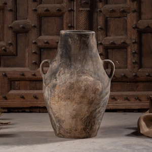 May include: A large, weathered clay vessel with a wide, rounded body and a narrow neck. The surface features a mottled, earthy color palette of dark charcoal, brown, and tan tones with a rough, uneven texture. It has two small, arched handles.
