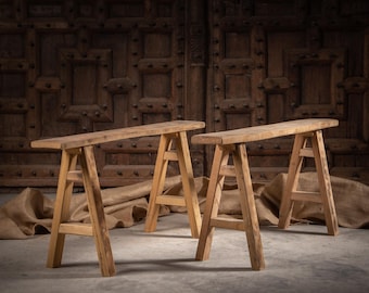 May include: Two light brown wooden trestle benches with a rectangular top and four angled legs connected by horizontal support bars. The benches feature a natural wood grain finish and a sturdy construction with a simple, functional design.