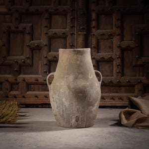 May include: A large, weathered, light brown clay vessel with a wide, rounded body and two small, curved handles on the sides. The surface has a matte, textured finish with subtle variations in tone and signs of age, featuring a wide, open top.
