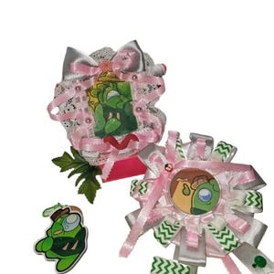 May include: Two decorative rosettes and a small acrylic charm featuring a green cartoon character. The rosettes are adorned with pink and white ribbons, lace, and small beads. The charm is a die-cut green figure with a brown hat and white visor.