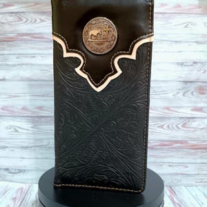 May include: A dark brown leather long wallet featuring an intricate floral embossed pattern and a smooth top section. A circular silver-toned metal concho is centered on the upper portion, displaying a raised relief of a cross and an elephant.