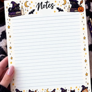 Puede incluir: Una hoja rectangular de papel rayado con la palabra Notes impresa en la parte superior. El borde presenta ilustraciones de gatos negros con sombreros de bruja, calabazas, lunas crecientes, estrellas, murciélagos, velas y un fantasma sobre un fondo blanco.