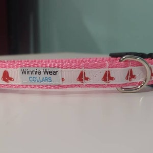 May include: Pink dog collar with a white ribbon featuring red roller skate graphics. The collar has a black buckle and a silver metal D-ring. The text "Winnie Wear COLLARS" is printed on the white ribbon.