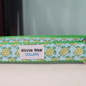 May include: A light blue dog collar with a pattern of yellow turtles. The collar has a green border and a metal D-ring. The label reads "Winnie Wear COLLARS".
