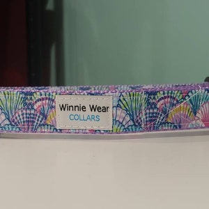 May include: A purple dog collar with a white background and a colorful seashell pattern. The collar has a silver metal buckle and a label that reads "Winnie Wear COLLARS".