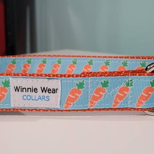 May include: A blue and orange dog collar with a repeating pattern of orange carrots on a light blue background. The collar has a silver D-ring and a black buckle. The collar has a white label that reads "Winnie Wear COLLARS".