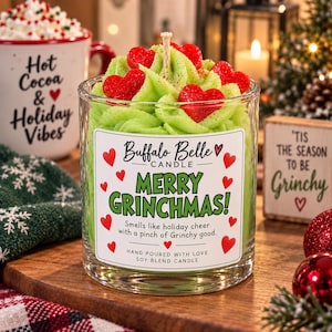 Puede incluir: Una vela en vaso de cristal transparente con cera verde moldeada como glaseado, decorada con pequeños corazones de cera roja y una mecha central. La etiqueta dice Buffalo Belle CANDLE MERRY GRINCHMAS! Smells like holiday cheer with a pinch of Grinchy good. HAND POURED WITH LOVE SOY BLEND CANDLE.