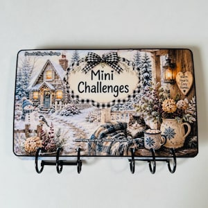 May include: A rectangular wall-mounted organizer featuring a winter scene with a snowy cottage, a snowman, and a cat. The text reads "Peaceful Kingdom Savings", "Mini Challenges", and "Warm Hearts Cozy Homes". It includes six black metal hooks.