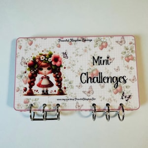 May include: A rectangular card with a strawberry and butterfly pattern featuring a cartoon girl with braided hair. The text reads Peaceful Kingdom Savings, Mini Challenges, and www.etsy.com/shop/PeacefulKingdomArt. The card has five metal binder rings.