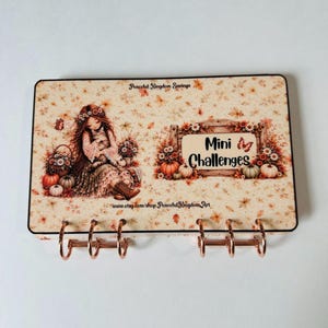 May include: A rectangular binder cover featuring a cream background with autumn leaves, pumpkins, and a girl holding a cat. The text reads "Peaceful Kingdom Savings" and "Mini Challenges" with a butterfly graphic. It has six rose gold metal rings.