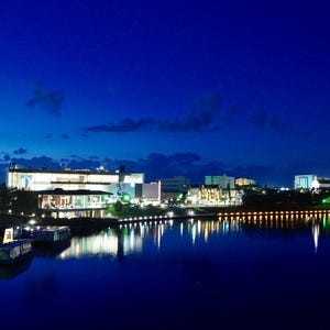 以下が含まれることがあります： 川岸に沿ってライトアップされた建物と、水面に停泊する数隻のボートが並ぶ夜の街並み。暗い青色の空と、建物から放たれる明るい人工的な光が対照的で、その光が穏やかな川面に鮮明に反射している様子が写し出されている。