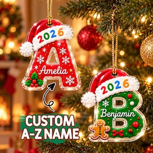 Pode incluir: Dois enfeites de Natal em forma de letra pendurados em cordões dourados. A letra A vermelha apresenta o nome Amelia, flocos de neve brancos e azevinho. A letra B verde apresenta o nome Benjamin, um boneco de gengibre e bagas vermelhas.