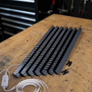May include: A set of six carbon fiber patterned triangular light channels arranged in parallel, accompanied by a coiled LED light strip with a white controller and two small black H-shaped plastic connectors for assembly.