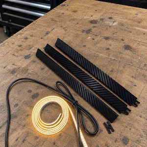 May include: Three black carbon fiber textured strips with small connector tabs, a coiled flexible LED light strip emitting a warm yellow glow, and a small black plastic connector piece arranged on a worn wooden workbench surface.