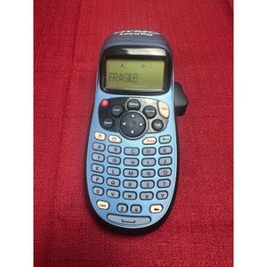 May include: A blue Dymo LetraTag label maker with a digital screen displaying the text FRAGILE. The device features a QWERTY keyboard with light blue circular buttons, a black navigation pad, and a top-mounted print button.