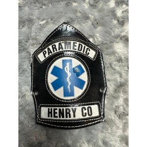 May include: A black leather shield-shaped patch with white stitching. It features a white banner at the top with the word PARAMEDIC in black, a central blue Star of Life emblem, and a white banner at the bottom with the text HENRY CO in black.
