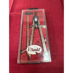 Peut inclure: Un compas de dessin en métal et une barre d'extension en métal séparée, logés dans un étui en plastique transparent avec un intérieur moulé rouge. Le compas présente un sommet en plastique noir avec la marque Utrecht imprimée en blanc. Un autocollant ovale blanc avec le mot Utrecht est apposé sur le couvercle.