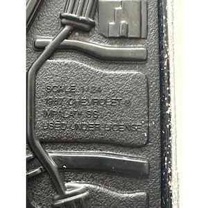 Könnte beinhalten: Eine schwarze, geformte Kunststoffbasis mit erhabenem Text, der SCALE 1/24 1967 CHEVROLET IMPALA SS USED UNDER LICENSE lautet. Die Oberfläche weist ein strukturiertes Finish mit geformten Strukturlinien und integrierten zylindrischen Komponenten auf.