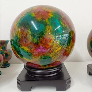 Puede incluir: Una esfera de piedra pulida con un patrón jaspeado de colores vibrantes, con remolinos de verde intenso, amarillo brillante, magenta y naranja. La esfera lisa y reflectante está montada sobre una base cuadrada de madera marrón oscuro.