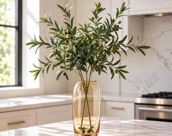 Könnte beinhalten: Eine hohe, zylindrische Vase aus bernsteinfarbenem Glas mit mehreren künstlichen Olivenzweigen. Die Zweige haben schmale, spitze grüne Blätter und kleine, runde grüne und dunkelbraune Oliven. Die Vase hat eine glatte, durchscheinende Oberfläche.