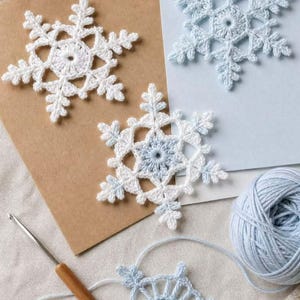 May include: Three finished crochet snowflakes in white and light blue, one partially completed snowflake attached to a ball of light blue yarn, and a wooden crochet hook. The snowflakes feature intricate, six-pointed geometric patterns.