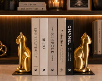May include: A pair of gold-toned cat-shaped bookends flanking a row of five books. The books are titled Live Beautifully, at 100, The Kinfolk Home, Minimalista, and Chanel Collections and Creations. The bookends feature a smooth, metallic finish.