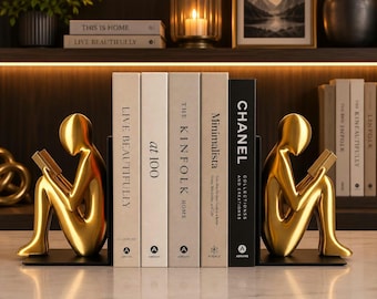 May include: Two gold-colored abstract human figures in a seated reading pose acting as bookends. Between them are five books with spines labeled: Live Beautifully, at 100, The Kinfolk Home, Minimalista, and Chanel Collections and Creations.
