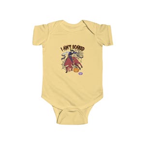 May include: A light yellow baby bodysuit featuring a graphic of a person riding a bull with the text I AIN'T SCARED. The design includes a spider web, bats, and a jack-o'-lantern pumpkin. The graphic is centered on the chest of the garment.