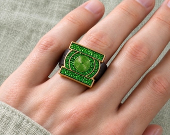 Puede incluir: Un anillo con un marco rectangular dorado, una incrustación verde texturizada y un emblema central en espiral verde. La banda del anillo es de color marrón oscuro y el diseño está inspirado en el símbolo del superhéroe Green Lantern.