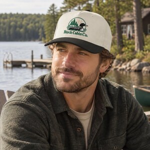 May include: A cream-coloured baseball cap with a black brim featuring a circular embroidered graphic of a loon on a lake with pine trees and the text Birch Cabin Co. in green lettering. The cap has a structured front panel and a button on top.