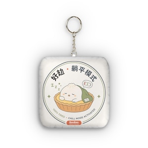 Puede incluir: Un llavero cuadrado con un personaje de bola de masa blanco descansando en una base de tarta amarilla. El diseño incluye texto en chino, un icono de batería baja y el texto en inglés 100% TIRED CHILL MODE ACTIVATED y BaoBao.