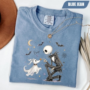 May include: A blue jean colored t-shirt featuring a graphic of a skeleton character in a pinstripe suit kneeling to touch the nose of a ghost dog. The design includes a crescent moon, bats, a spiderweb, stars, and the text Boop in black script.