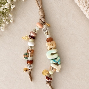 May include: A decorative keychain featuring two beige cord strands adorned with various beads, including clear, brown, and peach-colored spheres. One strand displays the letters R, U, T, Y in a mix of solid and plaid finishes, alongside small gold charms.