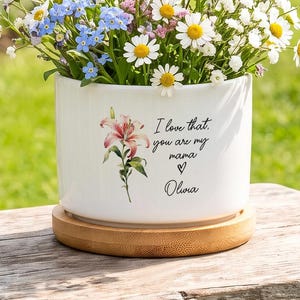 Puede incluir: Una maceta cilíndrica de cerámica blanca con base de bambú, que presenta una ilustración de un lirio rosa y el texto "I love that you are my mama" con un corazón y el nombre "Olivia", llena con un ramo de flores blancas, azules y amarillas.